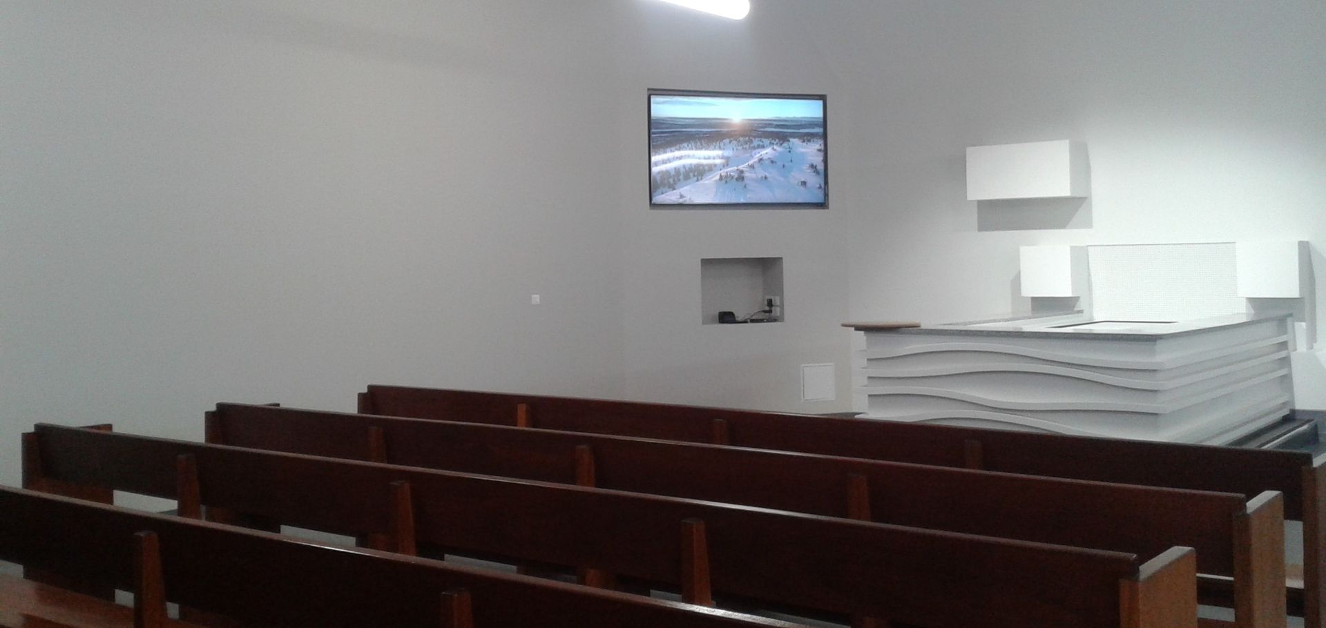 Salle de conférence avec des rangées de bancs en bois, un écran mural et des chaises blanches empilées.