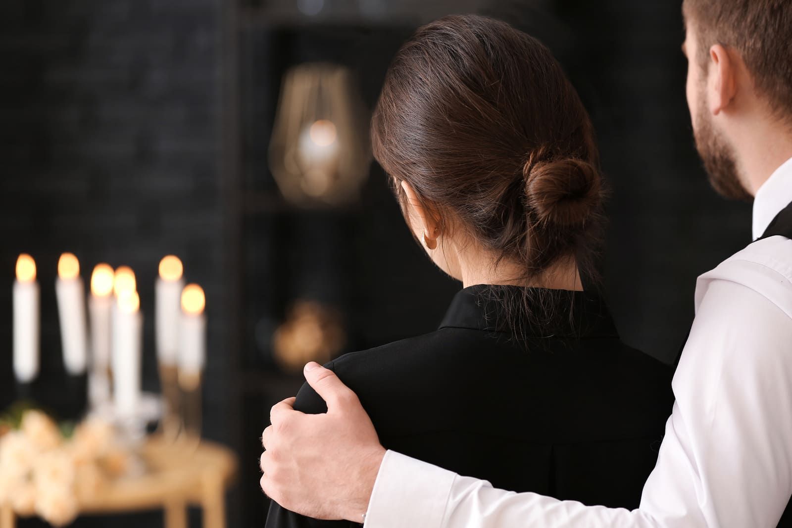 Deux personnes en tenue de soirée se tiennent côte à côte, l'une avec un bras autour de l'autre, dans un cadre faiblement éclairé à la bougie