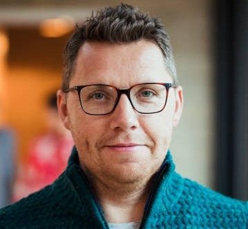 Un hombre que lleva gafas y un suéter verde mira a la cámara.
