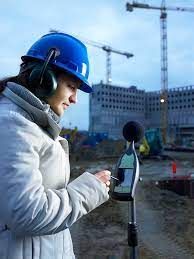 Una mujer que lleva casco y orejeras utiliza un medidor de sonido en un sitio de construcción.