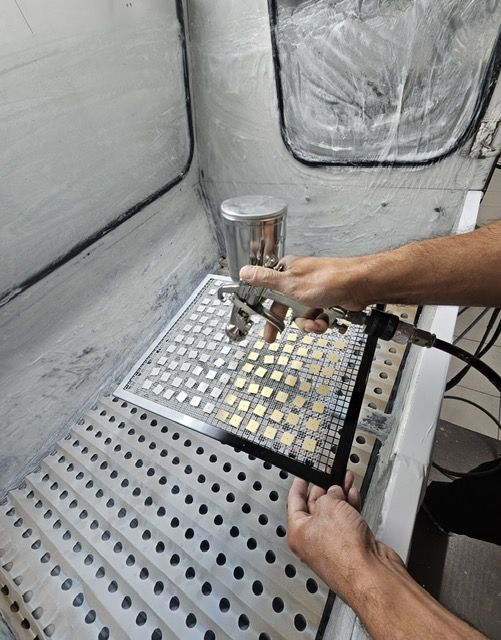 une main applique un vernis au pistolet sur des petites plaques dorées