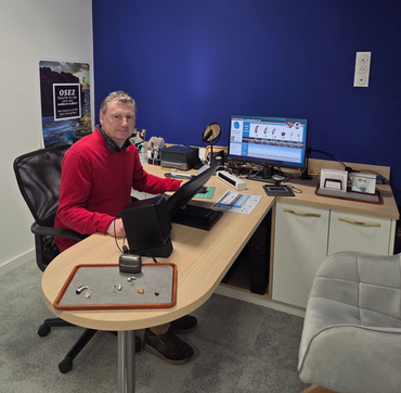 Un homme en pull rouge est assis à un bureau avec des écrans dans un bureau aux murs bleus.