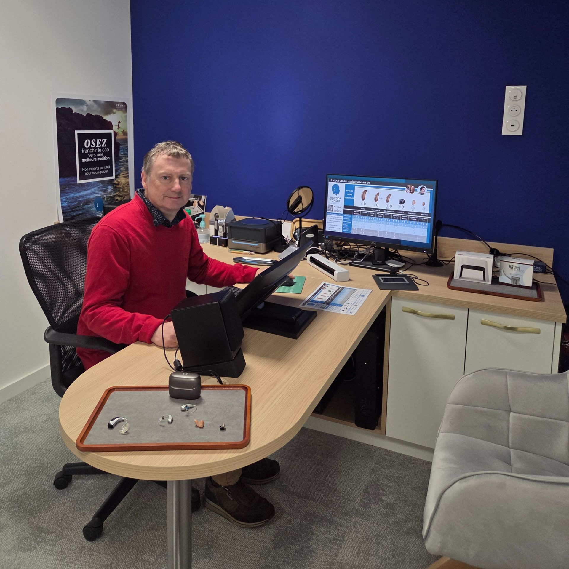 Un homme en pull rouge, assis à un bureau avec deux écrans, regarde la caméra.