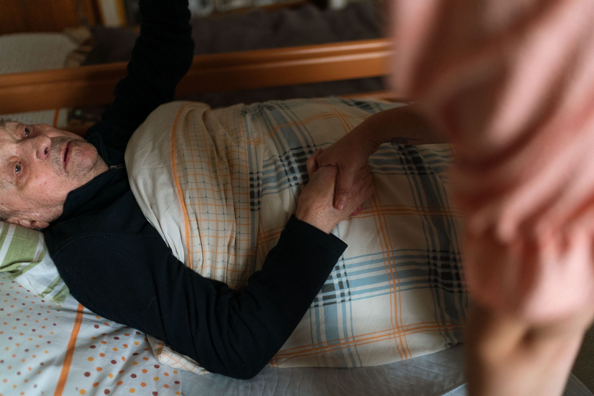 Personne âgée allongée dans son lit sous une couverture à carreaux, une main au premier plan.