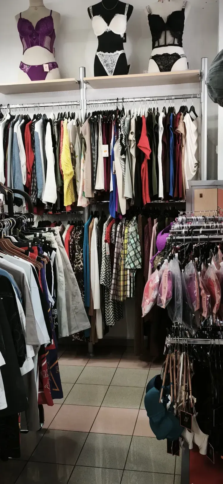 Pasillo de tienda de ropa con percheros llenos de ropa y sujetadores en el estante superior.