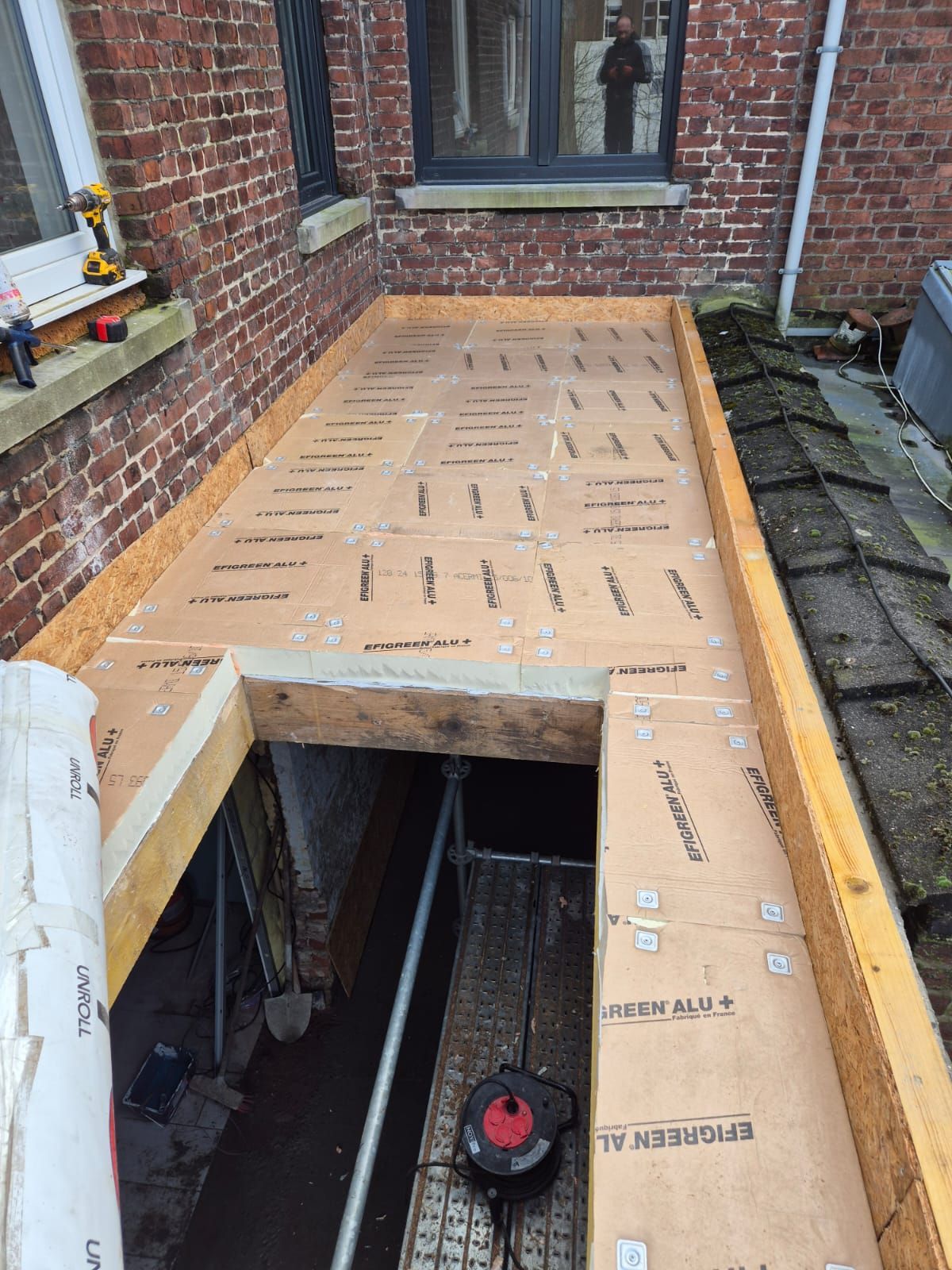 Chantier de construction avec charpente en bois apparente et toiture en panneaux de particules, à proximité d'un bâtiment en briques.