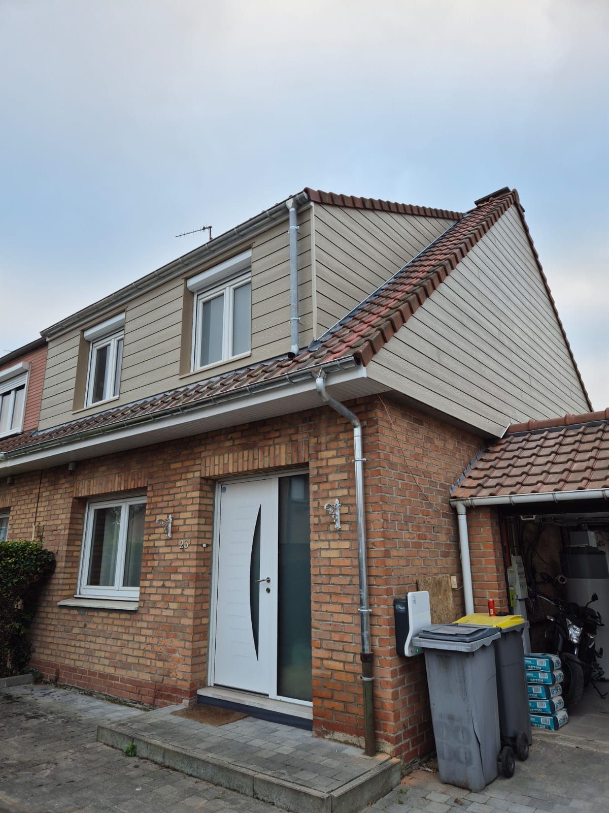 Maison mitoyenne en briques avec bardage beige, porte blanche et toit de tuiles rouges.