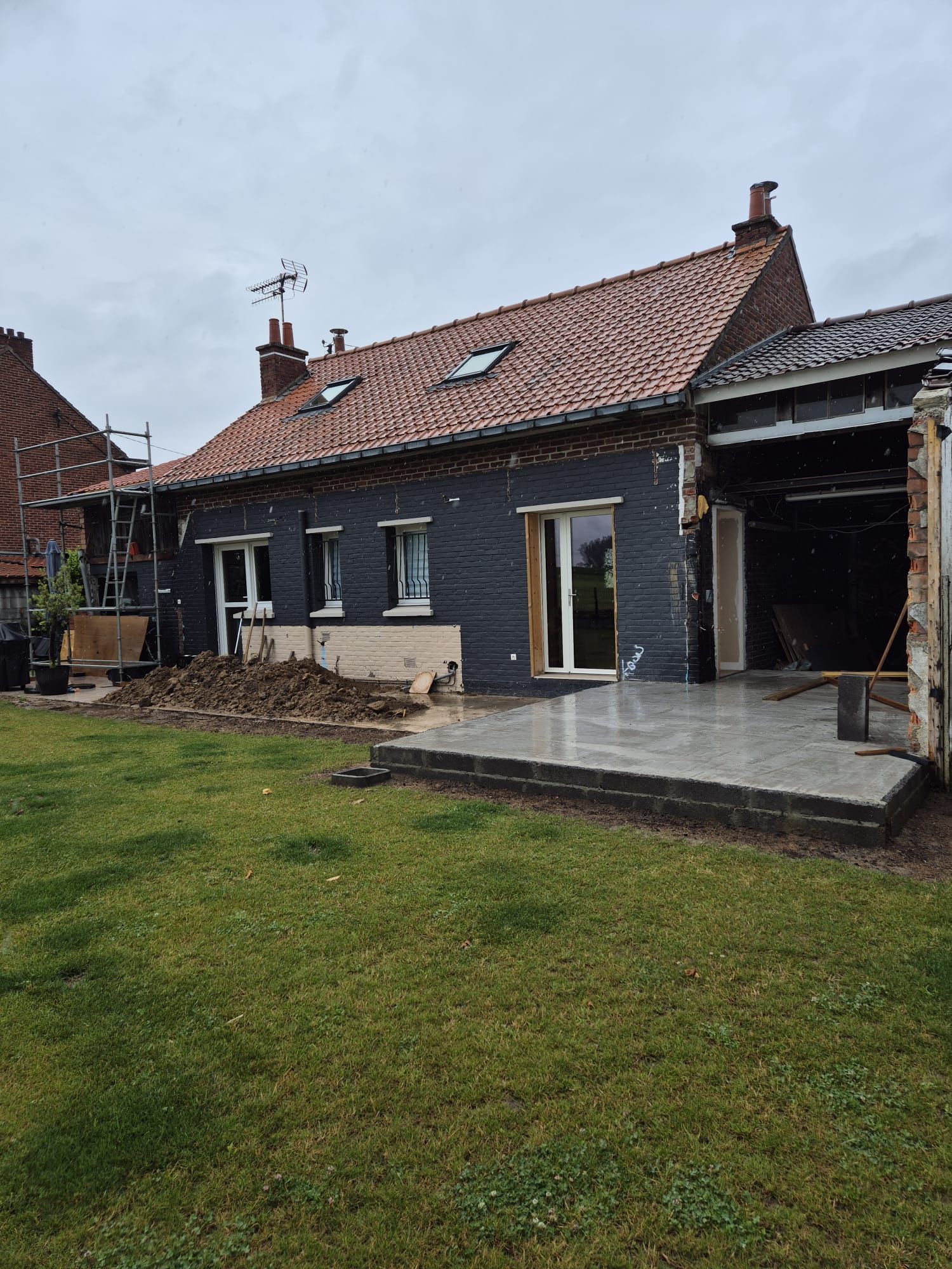 L'extérieur d'une maison en cours de rénovation : bardage foncé, toit de tuiles rouges et partie partiellement démolie.