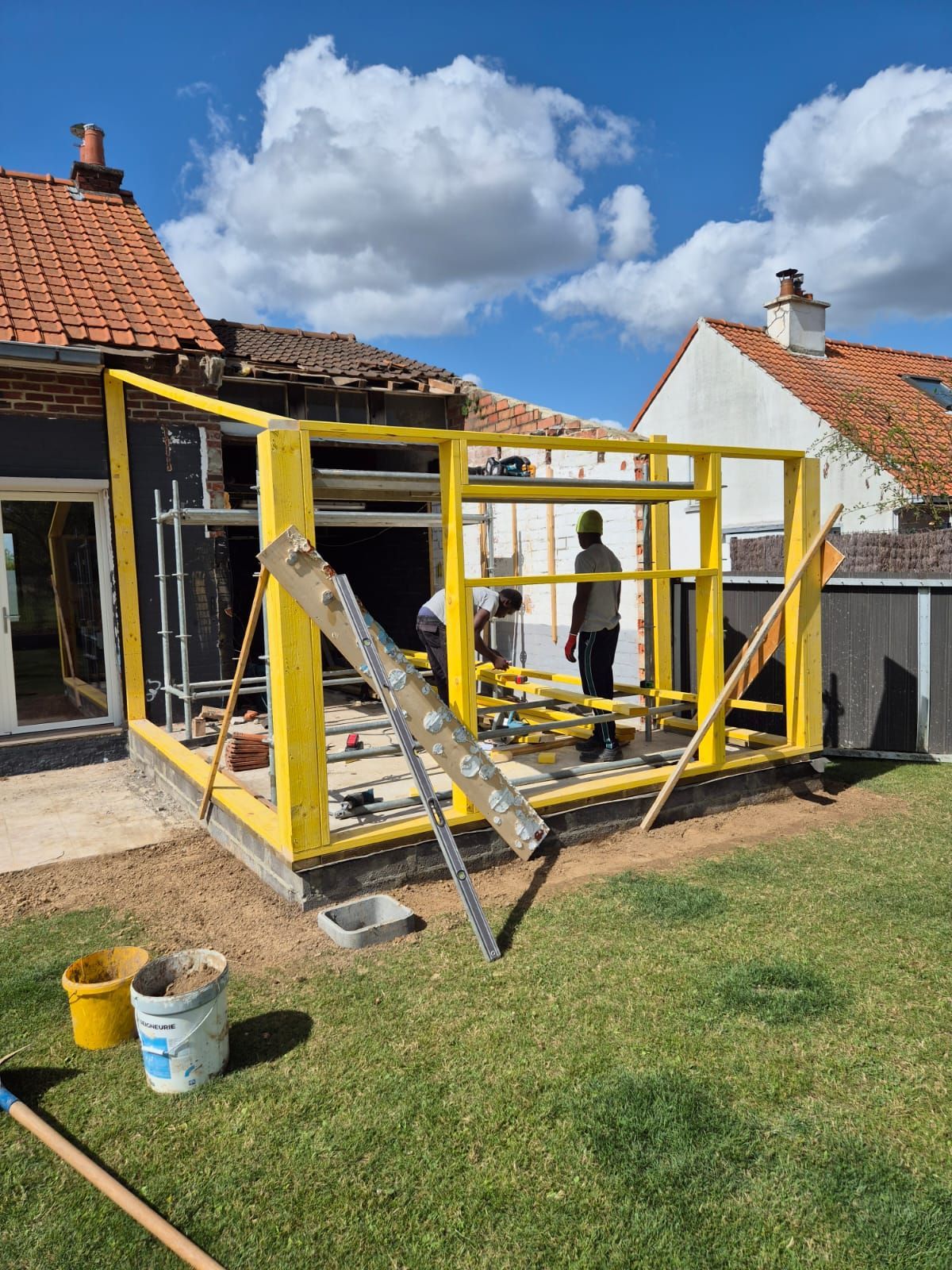 Structure à ossature jaune en construction, adossée à une maison ; deux personnes travaillent sur le chantier à l'extérieur par une journée ensoleillée.
