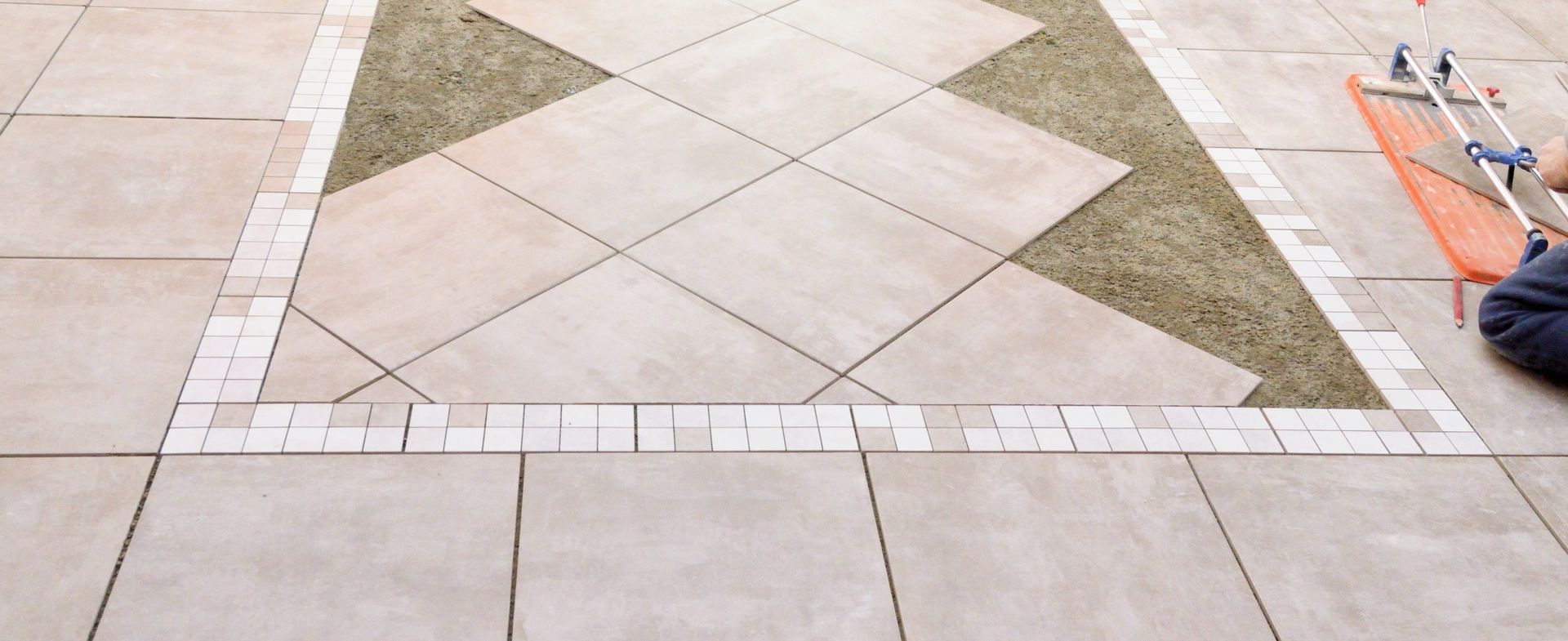 Terrasse aménagée avec carrelage à motifs géométriques, gazon et bordure, avec une personne utilisant un outil.