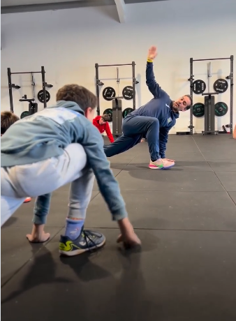 Personas estirando en el gimnasio; una persona con un brazo levantado. Equipo de pesas al fondo.
