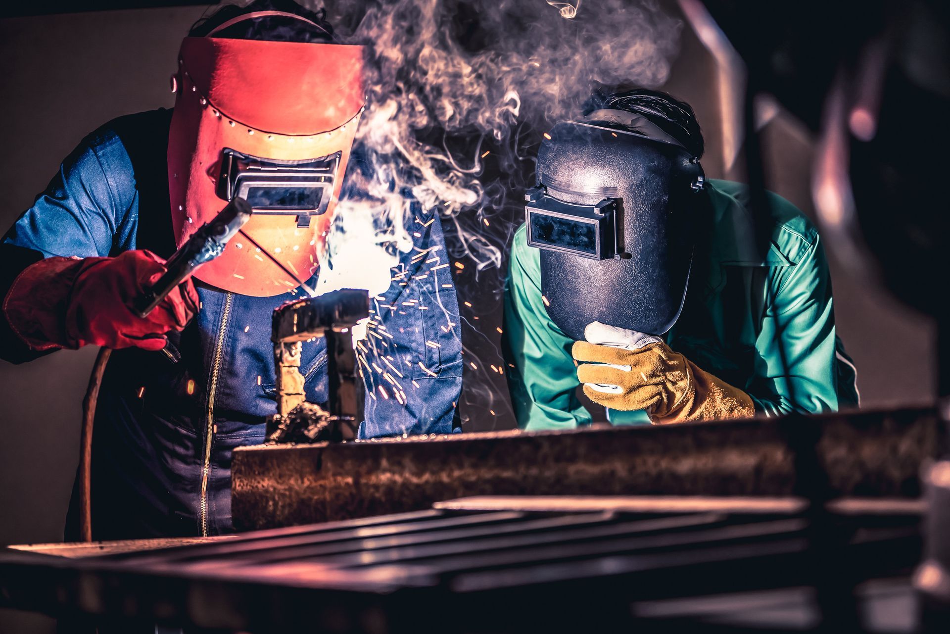 Dos soldadores con equipo de protección soldando metal en un taller; hay chispas y humo presentes.