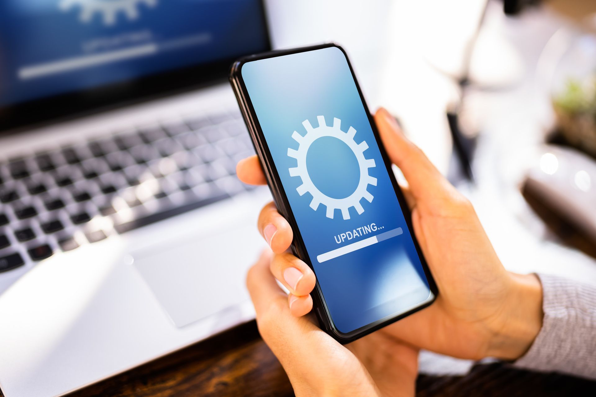 Persona sosteniendo un teléfono con un icono de engranaje en la pantalla, actualizándose. Portátil visible al fondo.