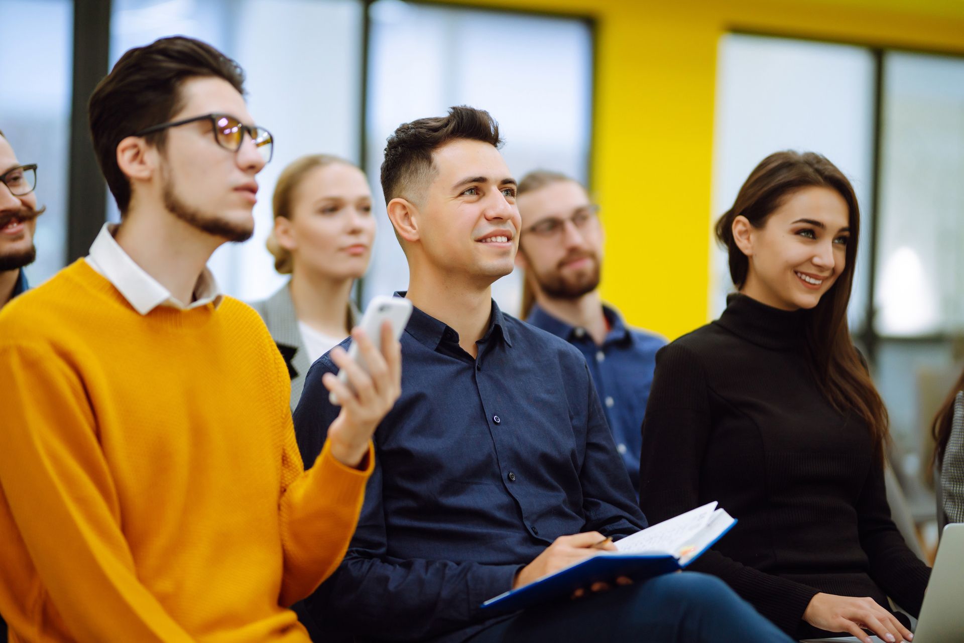 Grupo de personas diversas que asisten a una presentación, algunas sonriendo, en una sala bien iluminada.