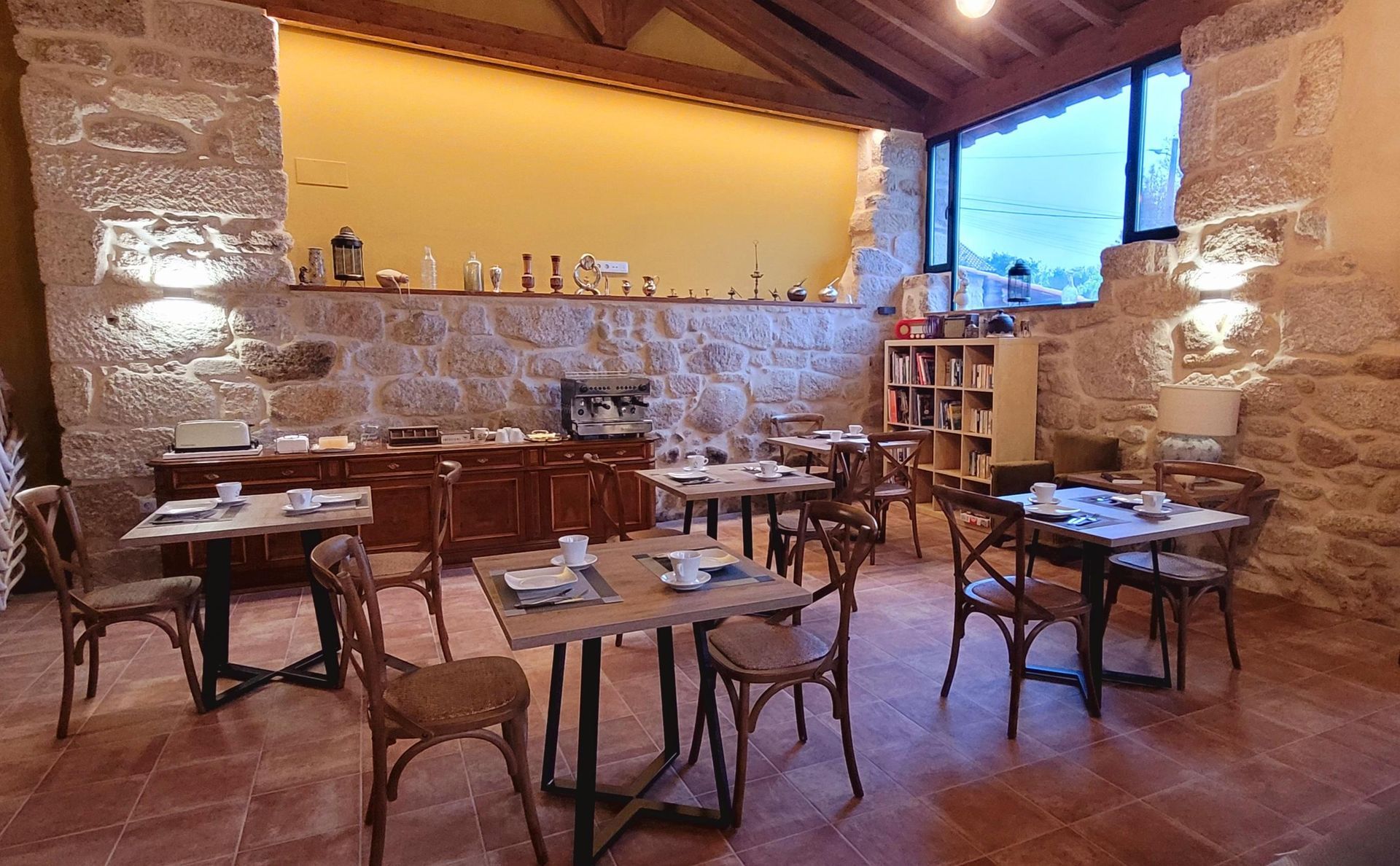 Salón de desayuno con paredes de piedra, mesas y sillas de madera, buffet y vista a la ventana.