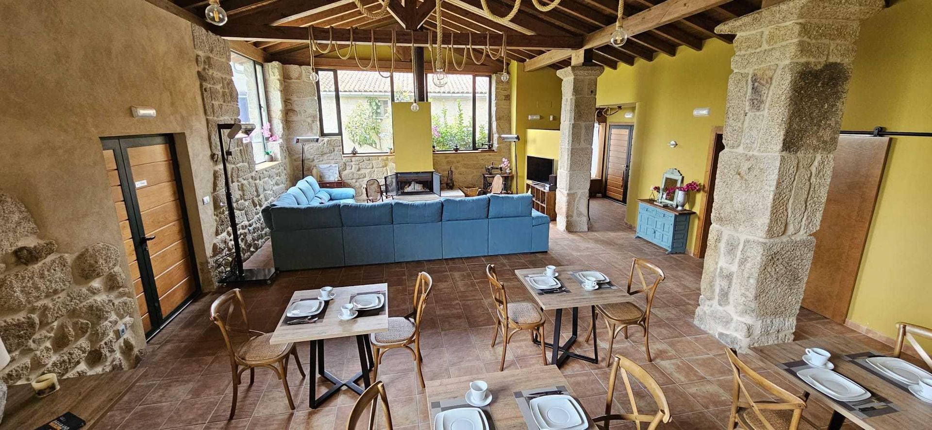 Interior de restaurante con mesas dispuestas para cenar, pilares de piedra y un sofá azul al fondo.