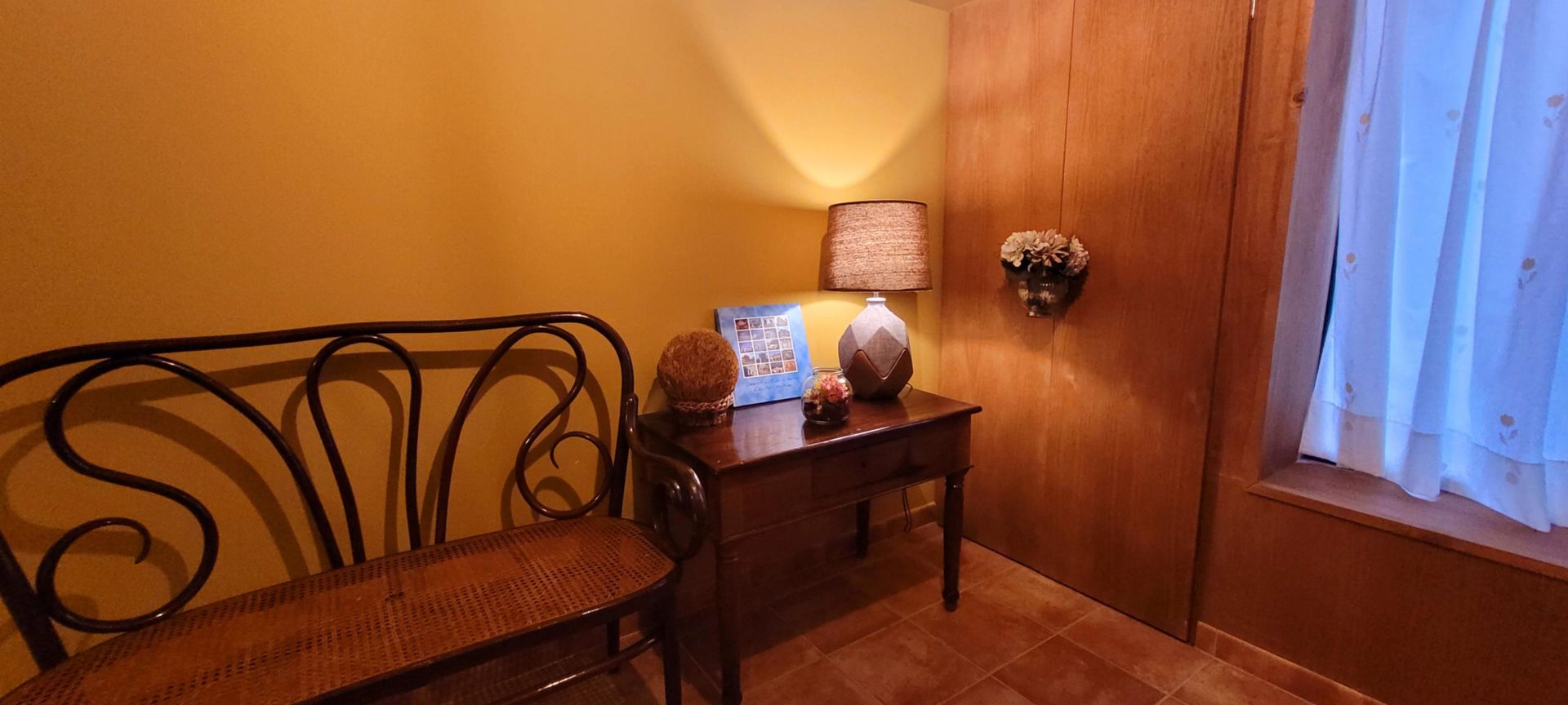 Un rincón acogedor con banco, mesa, lámpara y ventana con cortinas. Paredes de cálido amarillo y tonos madera.