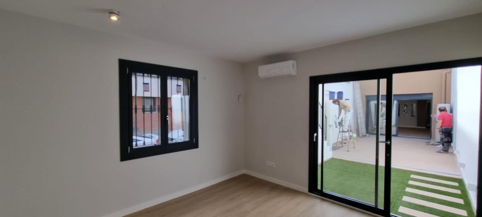 Vista interior con ventana y puerta corrediza de vidrio que da a un patio con césped. Aire acondicionado montado en la pared.