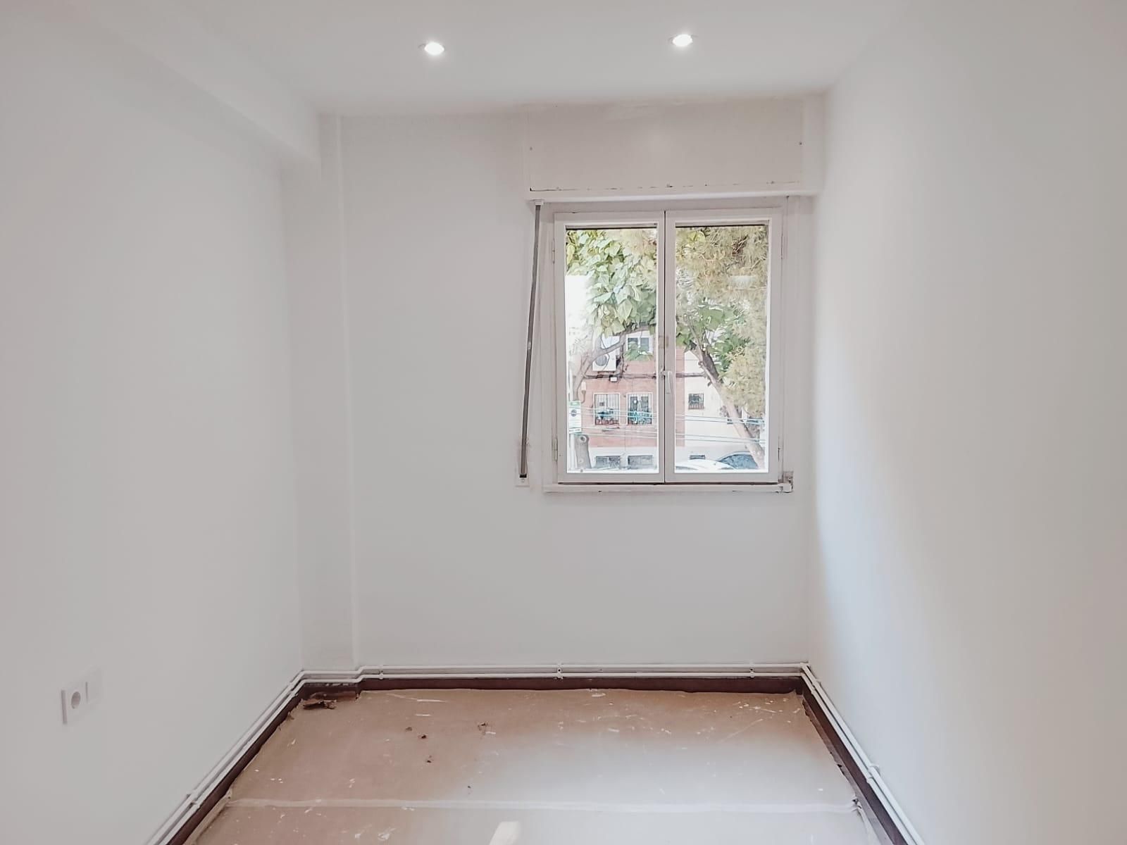 Habitación vacía de paredes blancas con una ventana, dos luces en el techo y pisos desnudos