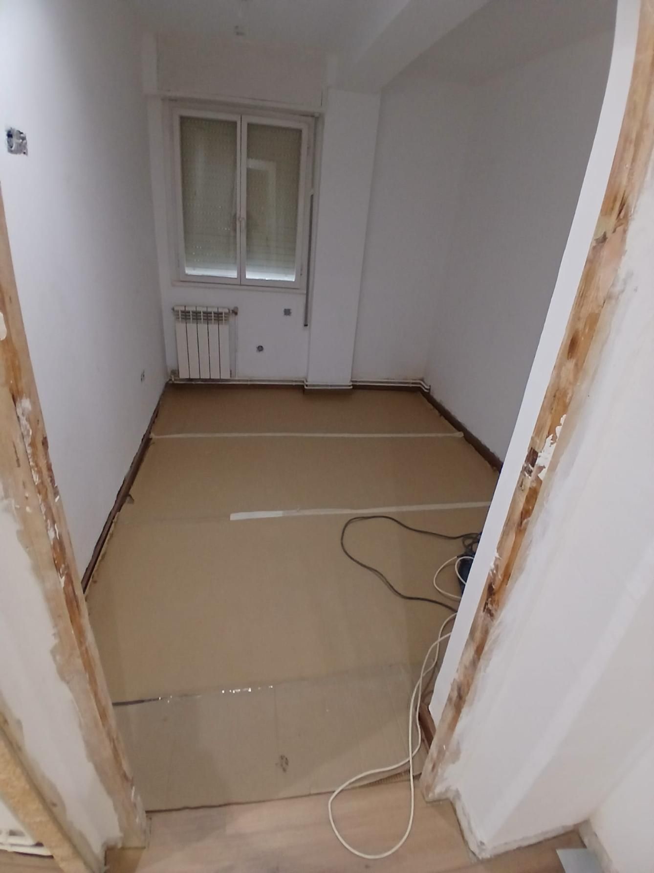 Habitación vacía con piso sin terminar, ventana, radiador y puerta expuesta.