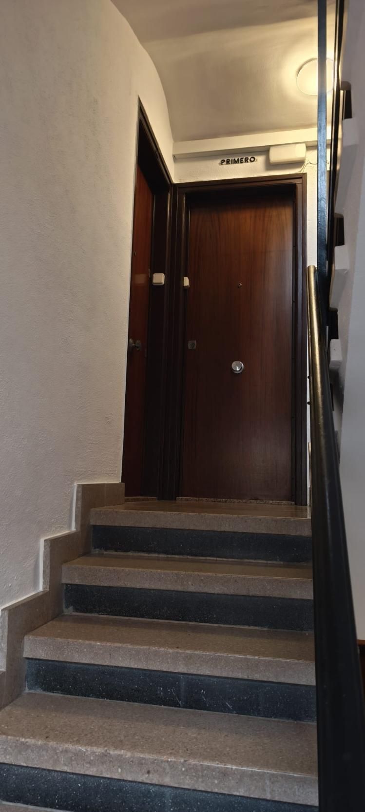 Escalera que sube a un pasillo con puerta de madera cerrada. Escalones grises y pared blanca.