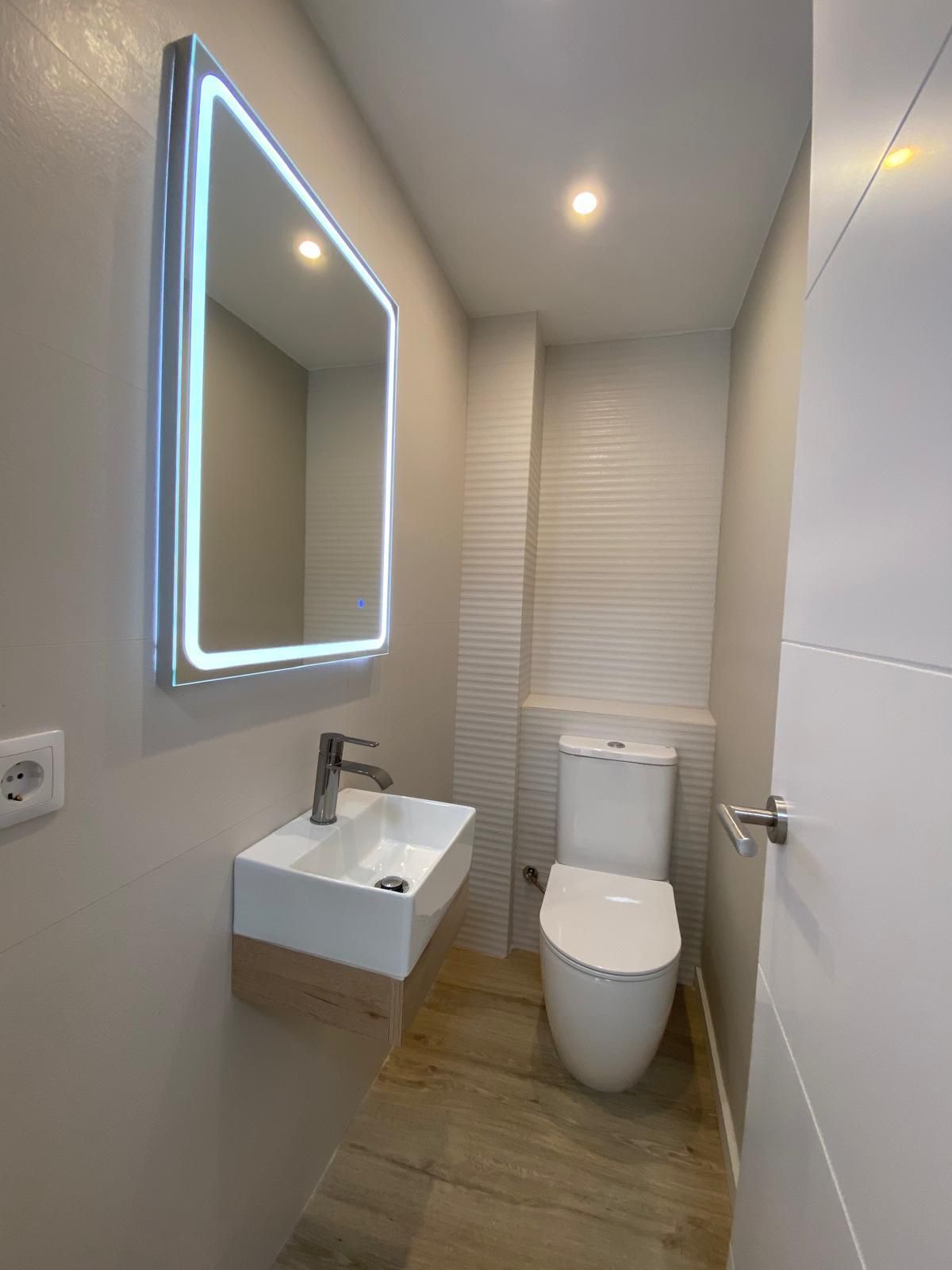 Tocador moderno con lavabo, inodoro y espejo blancos con iluminación LED. Paredes beige y suelo de baldosas imitación madera.