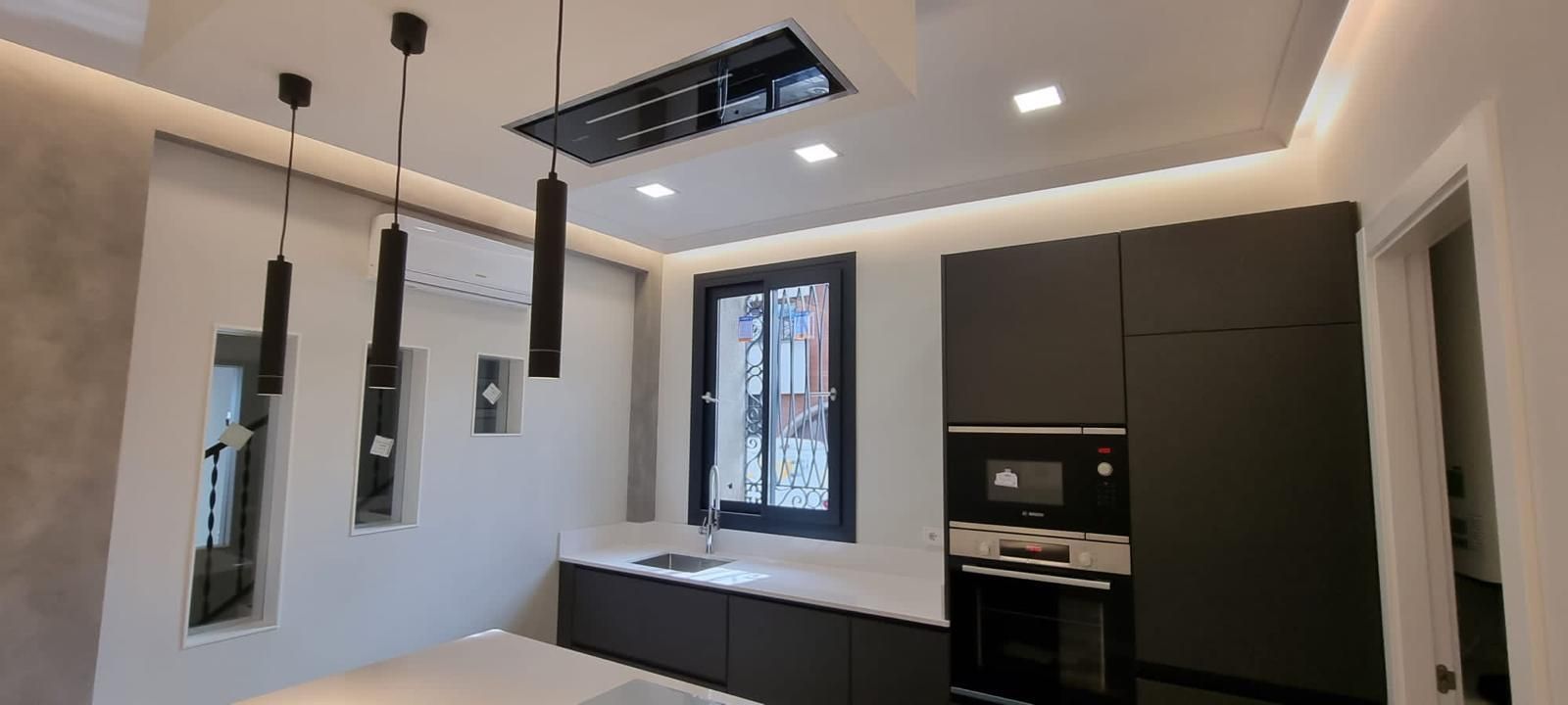 Cocina moderna con gabinetes oscuros, encimeras blancas y luces colgantes negras.