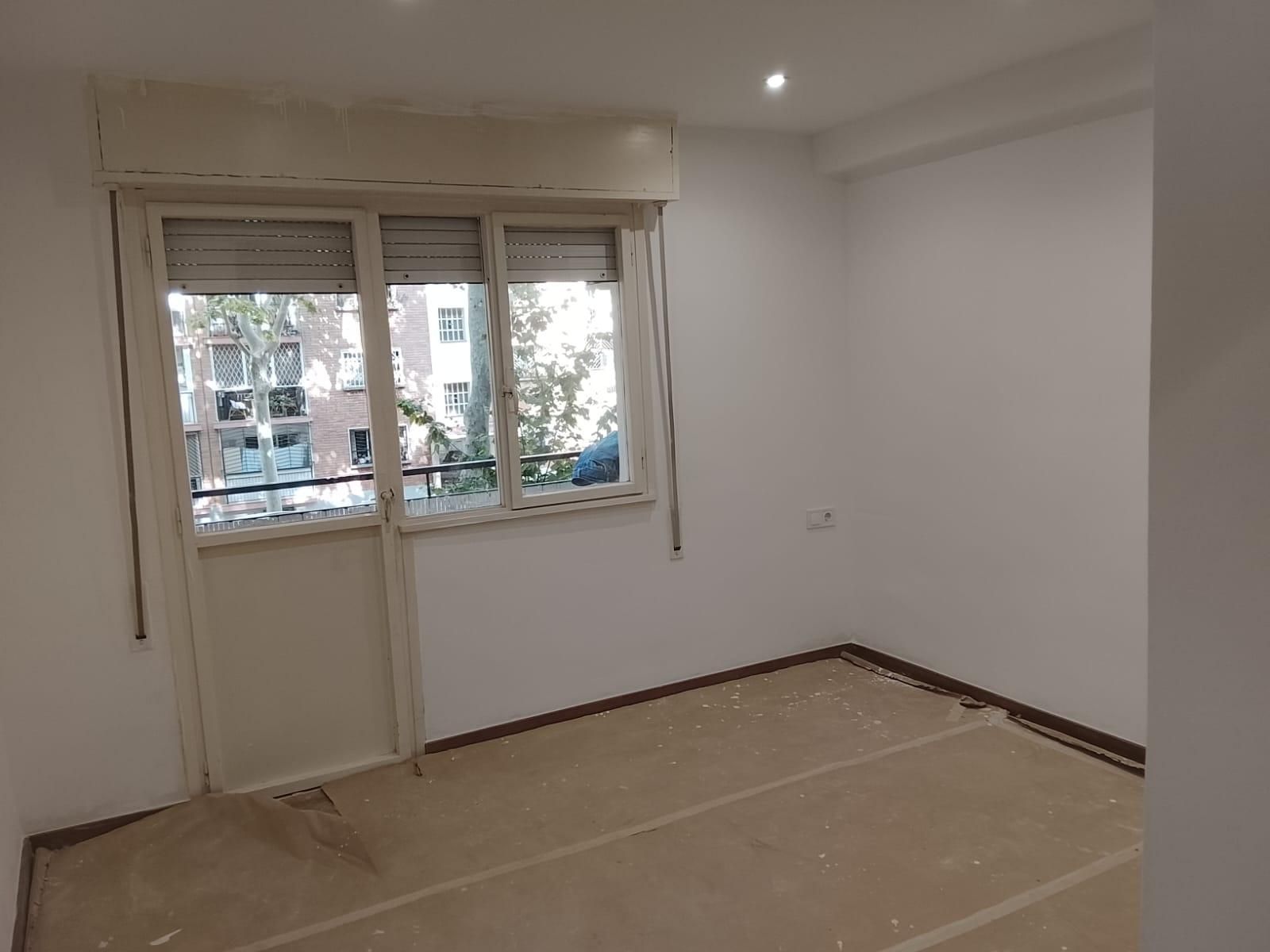 Habitación vacía con una ventana y una puerta cerrada. Las paredes son blancas, el suelo está tapizado