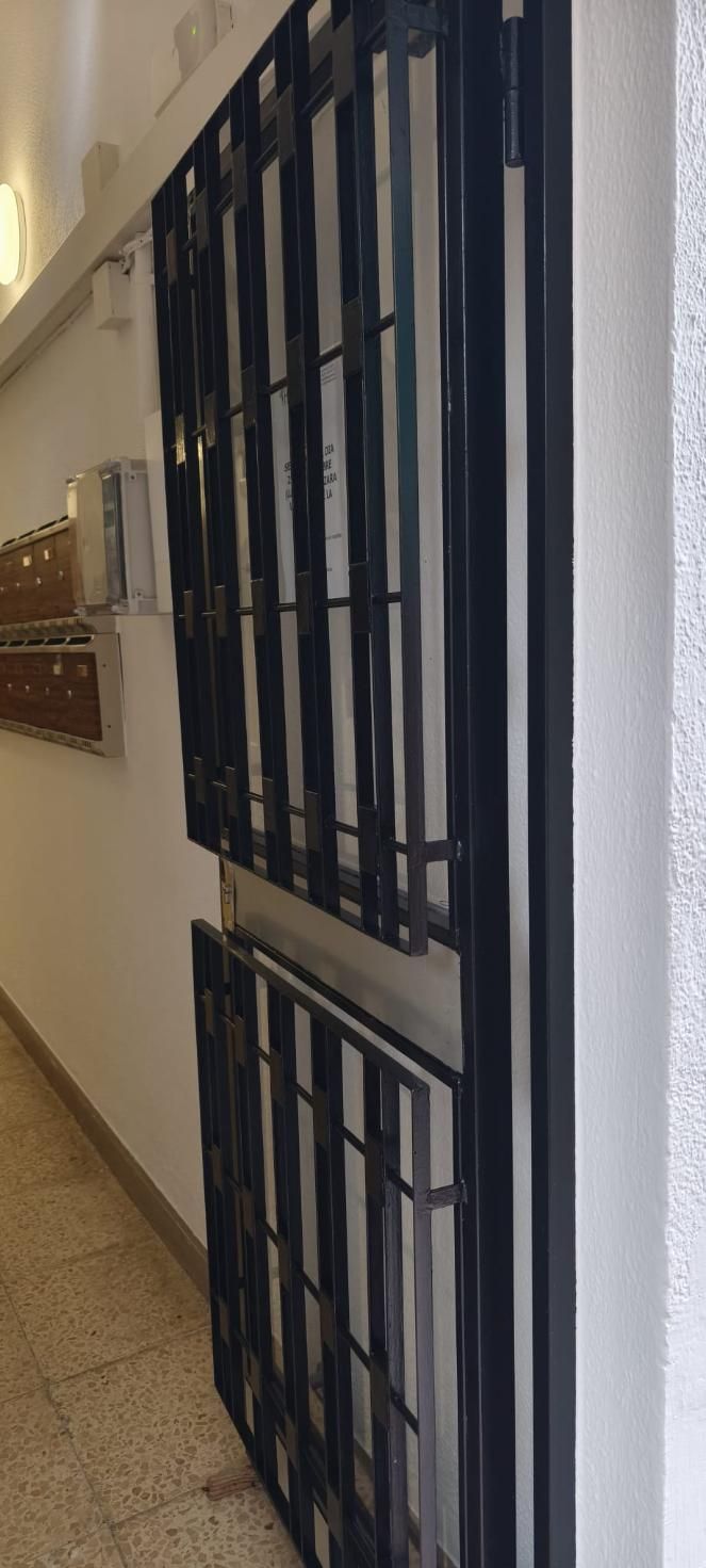 Una reja de seguridad metálica negra está abierta en un pasillo. Suelo de baldosas beige moteadas y paredes blancas.