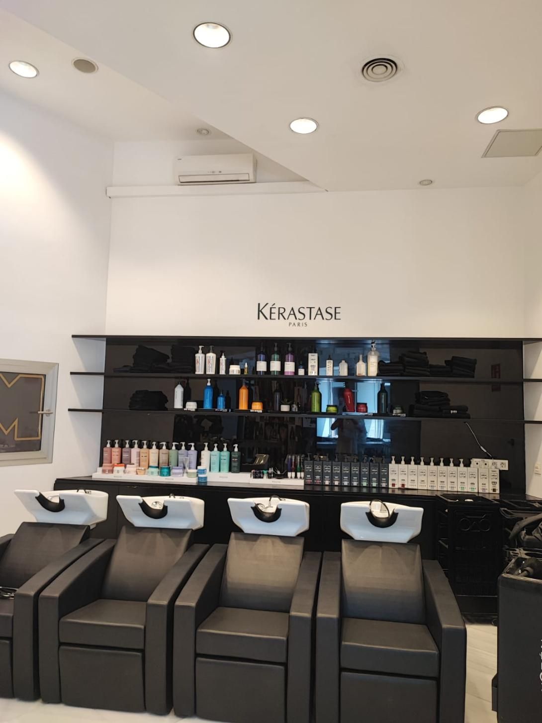Salón con sillas y lavabos negros, productos Kerastase en estantes e iluminación brillante.