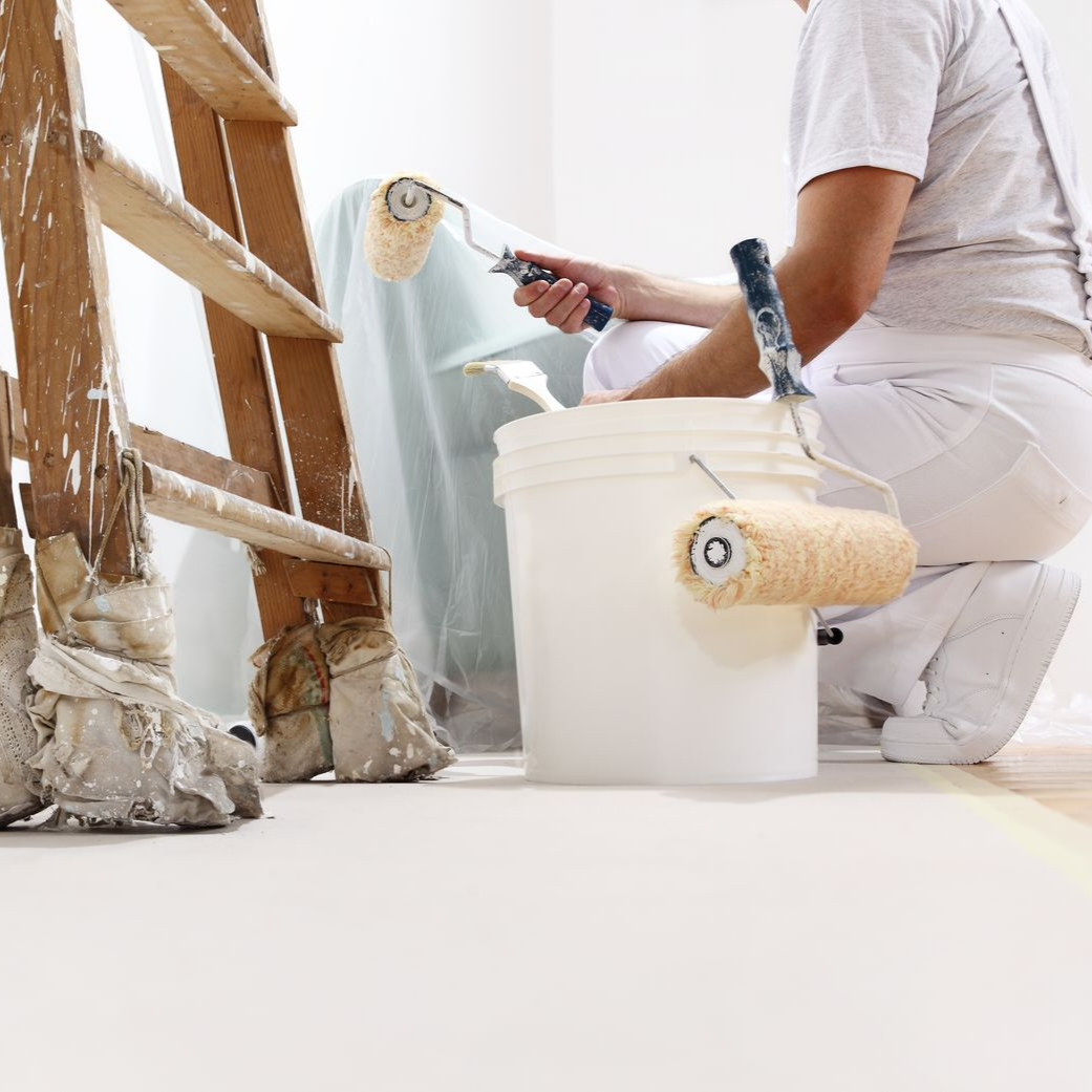 Un peintre en salopette blanche utilise un rouleau à peinture, une échelle et un seau de peinture dans une pièce claire.