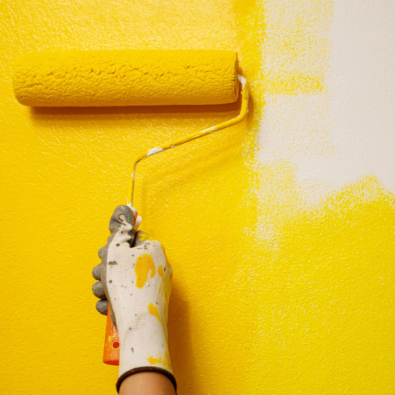 Une main peint un mur blanc avec un rouleau à peinture jaune ; une main gantée est visible.