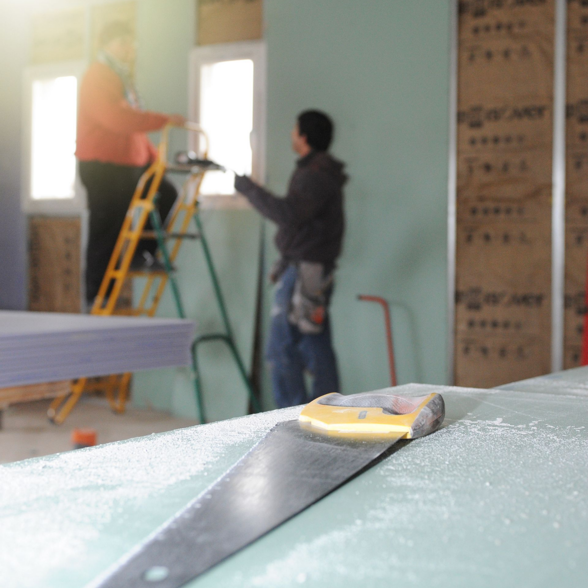 Des ouvriers du bâtiment installent des plaques de plâtre, l'un d'eux sur une échelle, scie au premier plan.