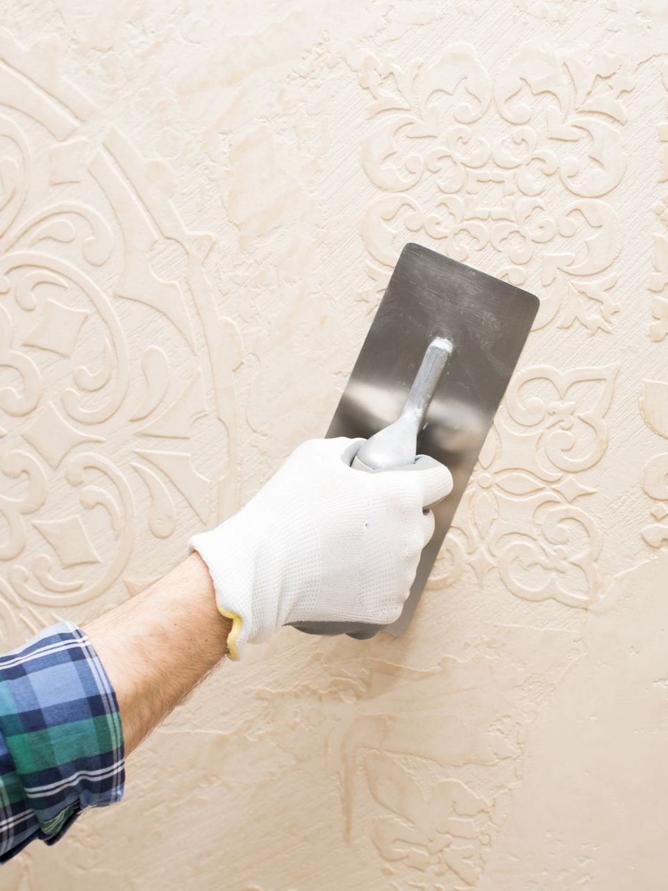 Une personne utilise une truelle pour appliquer un enduit à motifs floraux sur un mur, portant un gant et une chemise à carreaux.