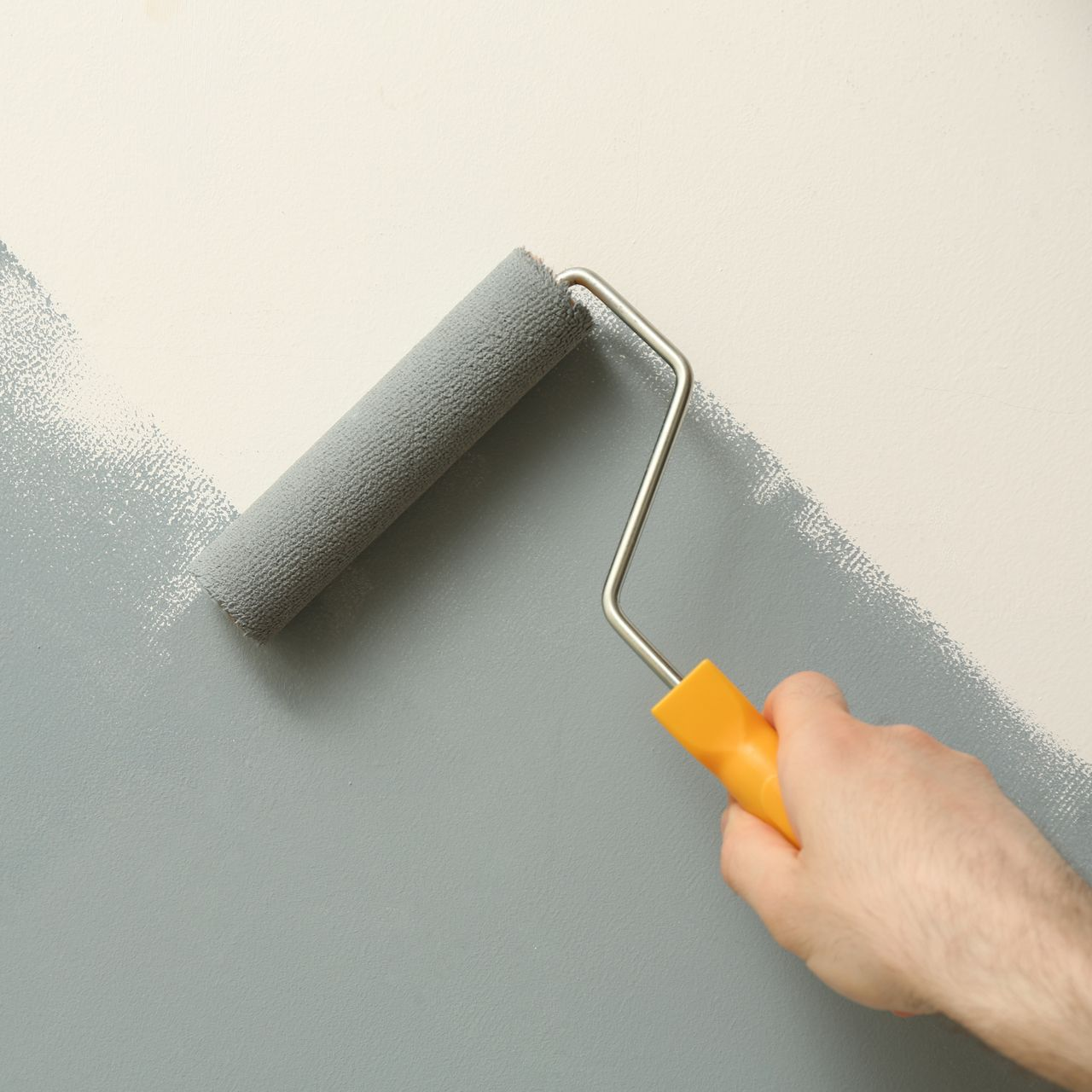 Main tenant un rouleau à peinture appliquant de la peinture grise sur un mur.