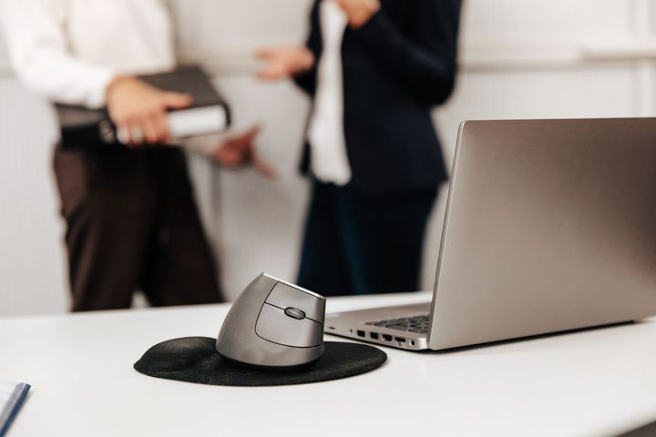 Zwei Personen in Anzügen arbeiten an einem Schreibtisch, eine schreibt, die andere benutzt einen Taschenrechner; Dokumente und Laptop sind vorhanden.