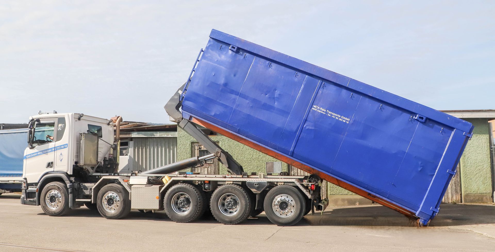 Ein blauer Industriecontainer, gefüllt mit Kies, vor einem grünen Gebäude.