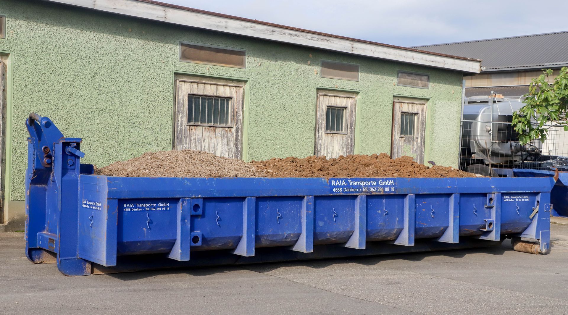 Ein blauer Müllcontainer vor einem grünen Gebäude mit zwei verwitterten Türen und einem kleinen Fenster.
