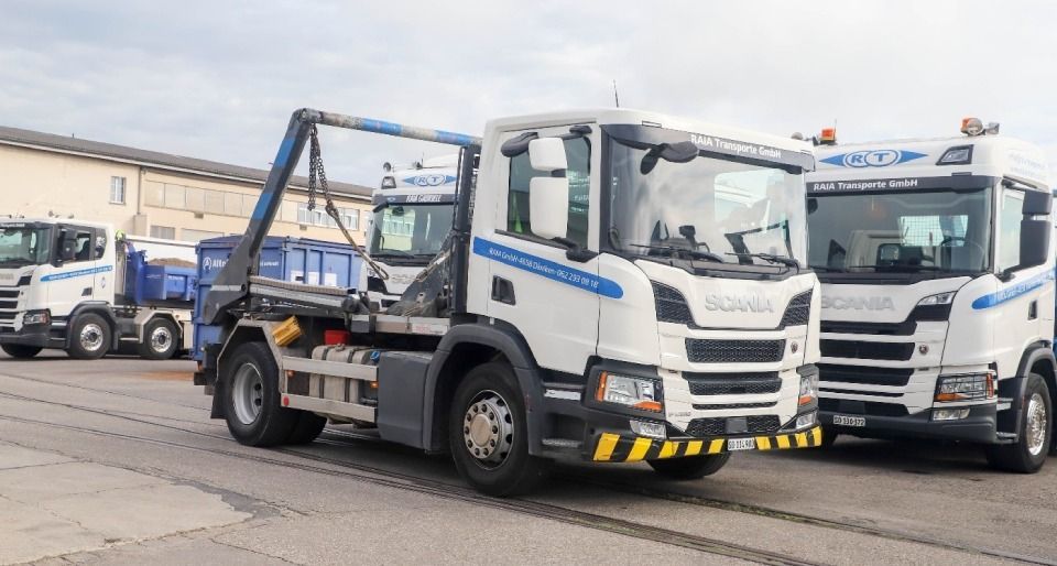 Weisse Scania-Lkw mit Abfallcontainern, geparkt im Freien unter bewölktem Himmel.