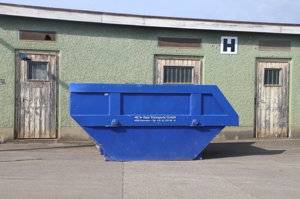 Ein blauer Müllcontainer vor einem grünen Gebäude mit zwei verwitterten Türen und einem kleinen Fenster.