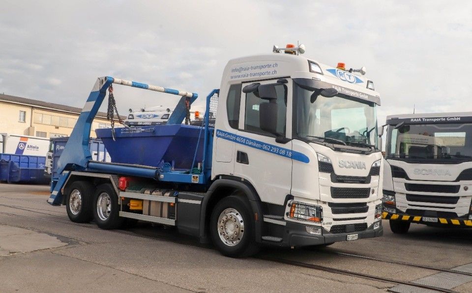 Ein weiss-blauer Scania-Abrollkipper mit einem Container steht im Freien neben einem anderen Lkw.