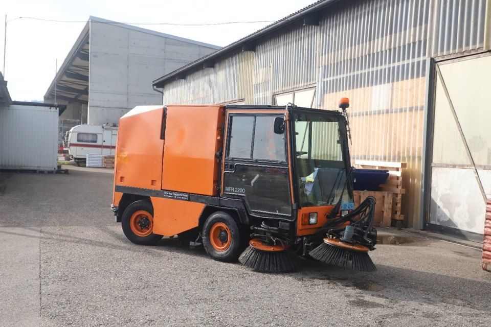 Eine orangefarbene Strassenkehrmaschine parkt auf einer gepflasterten Strasse in der Nähe von Gebäuden.