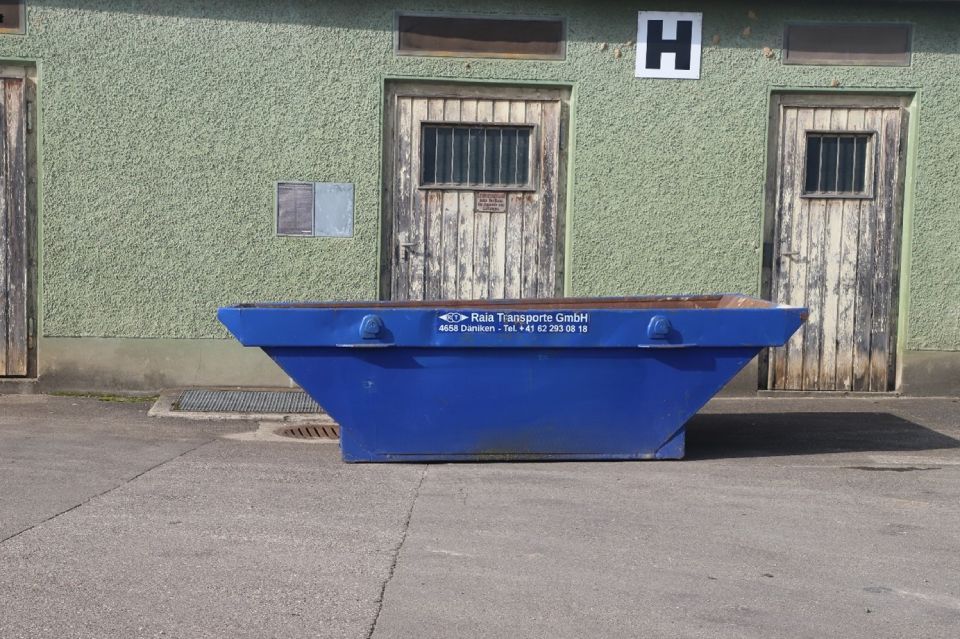 Ein blauer Müllcontainer vor einem verwitterten grünen Gebäude mit Holztüren.