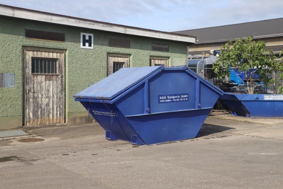 Ein blauer Industriecontainer vor einem grünen Gebäude mit einem „H“ über der Tür an einem sonnigen Tag.