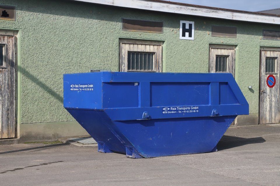 Ein blauer Müllcontainer vor einem verwitterten grünen Gebäude mit Holztüren und -fenstern.