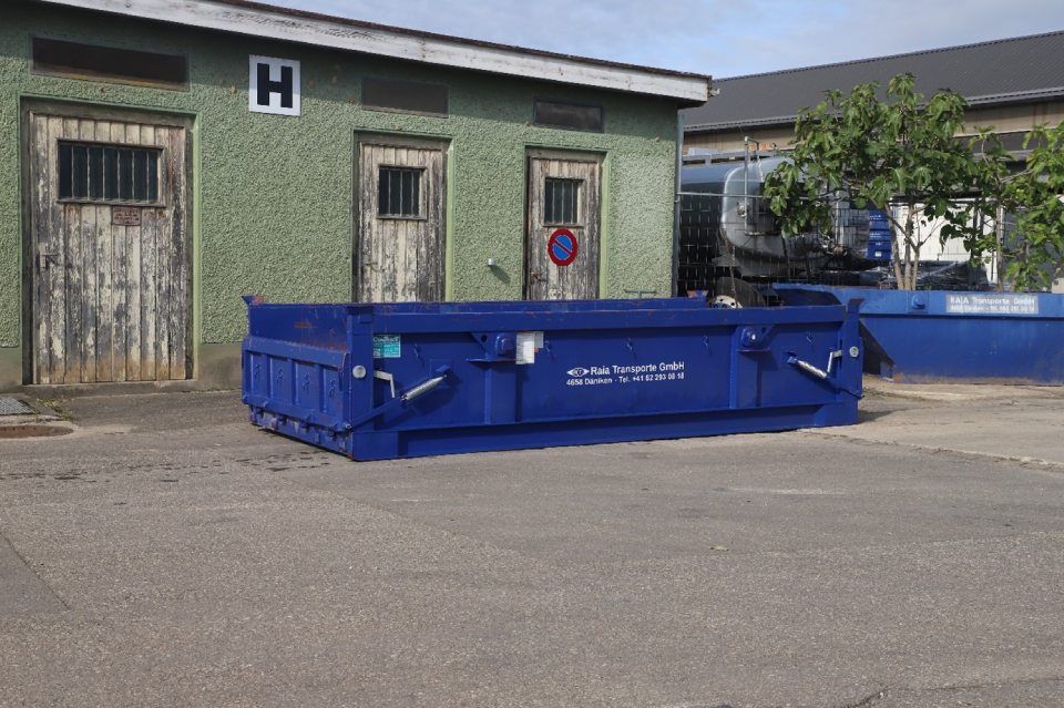 Ein blauer Müllcontainer vor einem grünen Gebäude mit drei Holztüren.