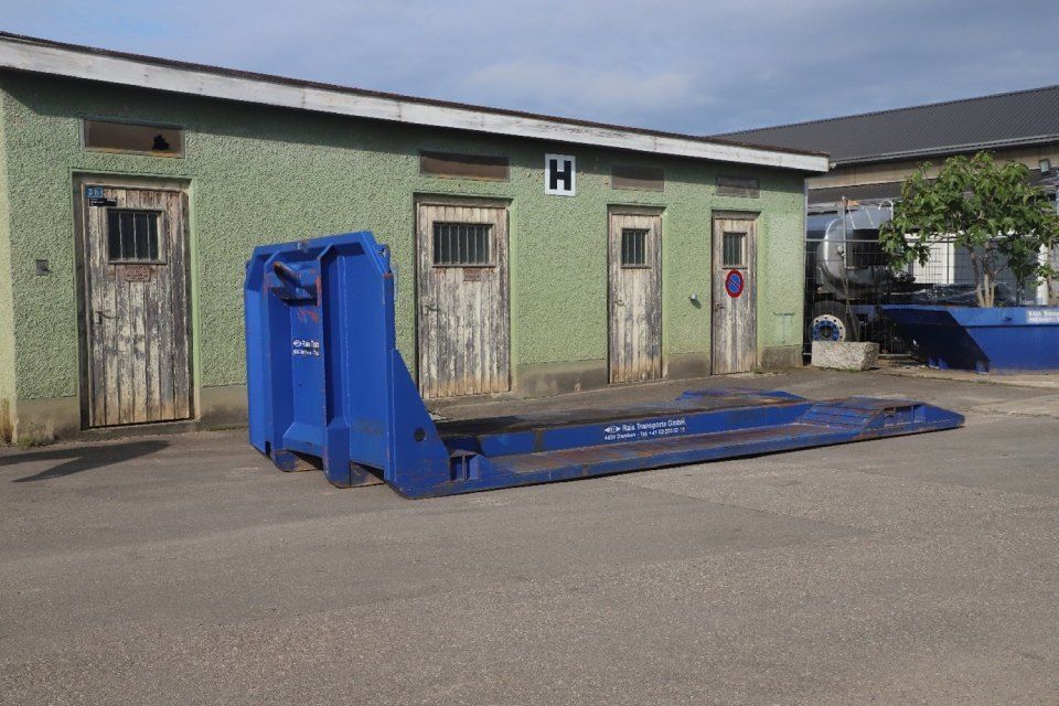 Ein blauer Anhänger parkte vor einem grünen Gebäude mit Holztüren und bewölktem Himmel.