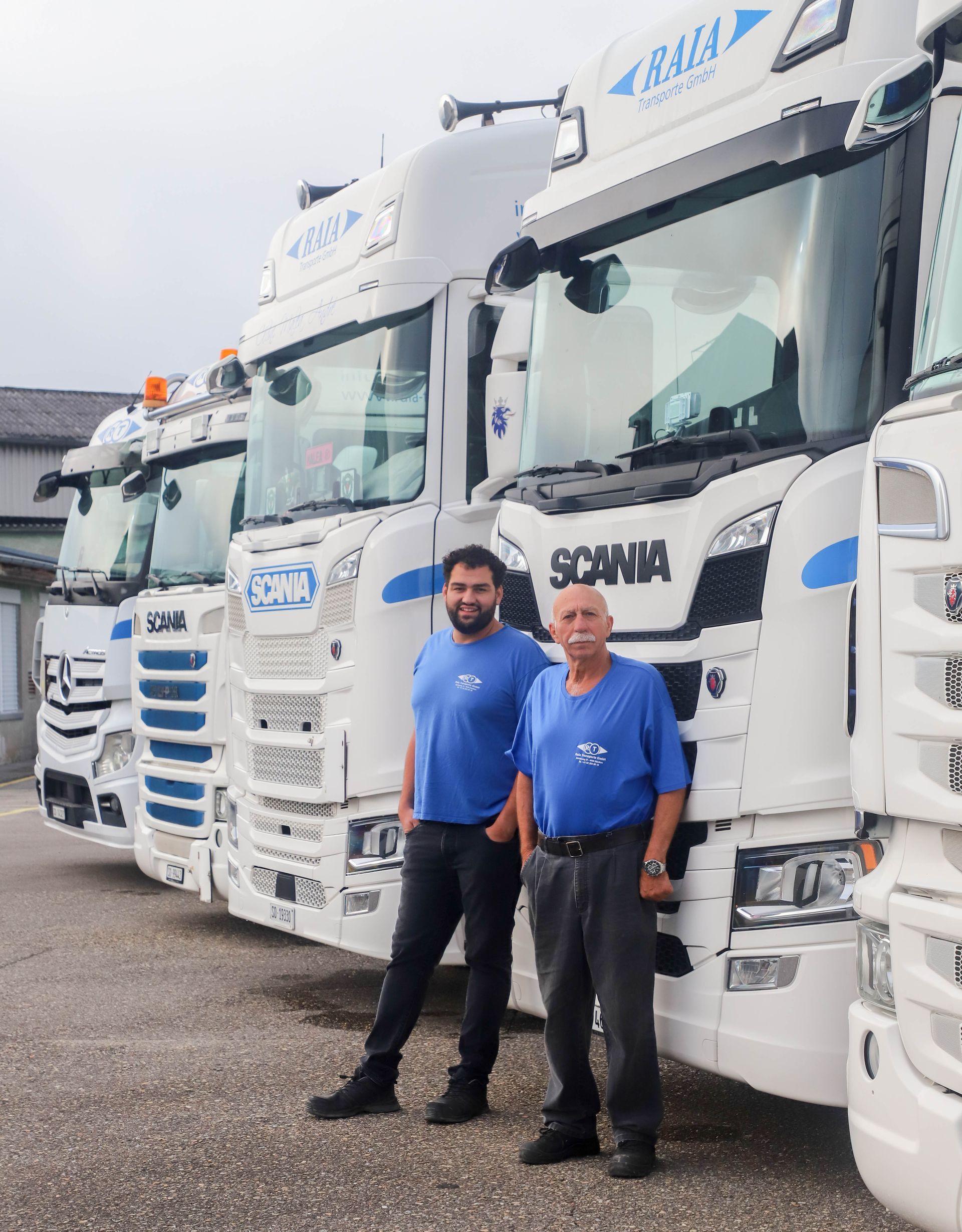 Eine Gruppe weisser Lastwagen mit Fahrern posiert an einem bewölkten Tag vor einer Fabrik.