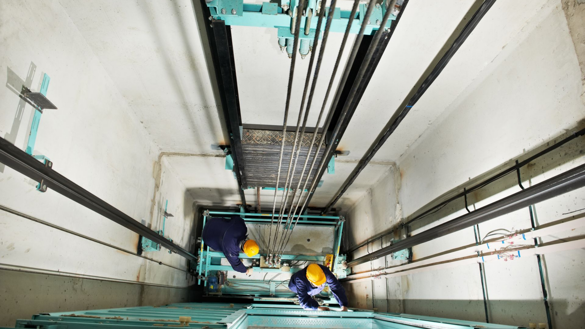 Des ouvriers portant des casques bleus et jaunes inspectent les machines d'ascenseur à l'intérieur d'une cage.