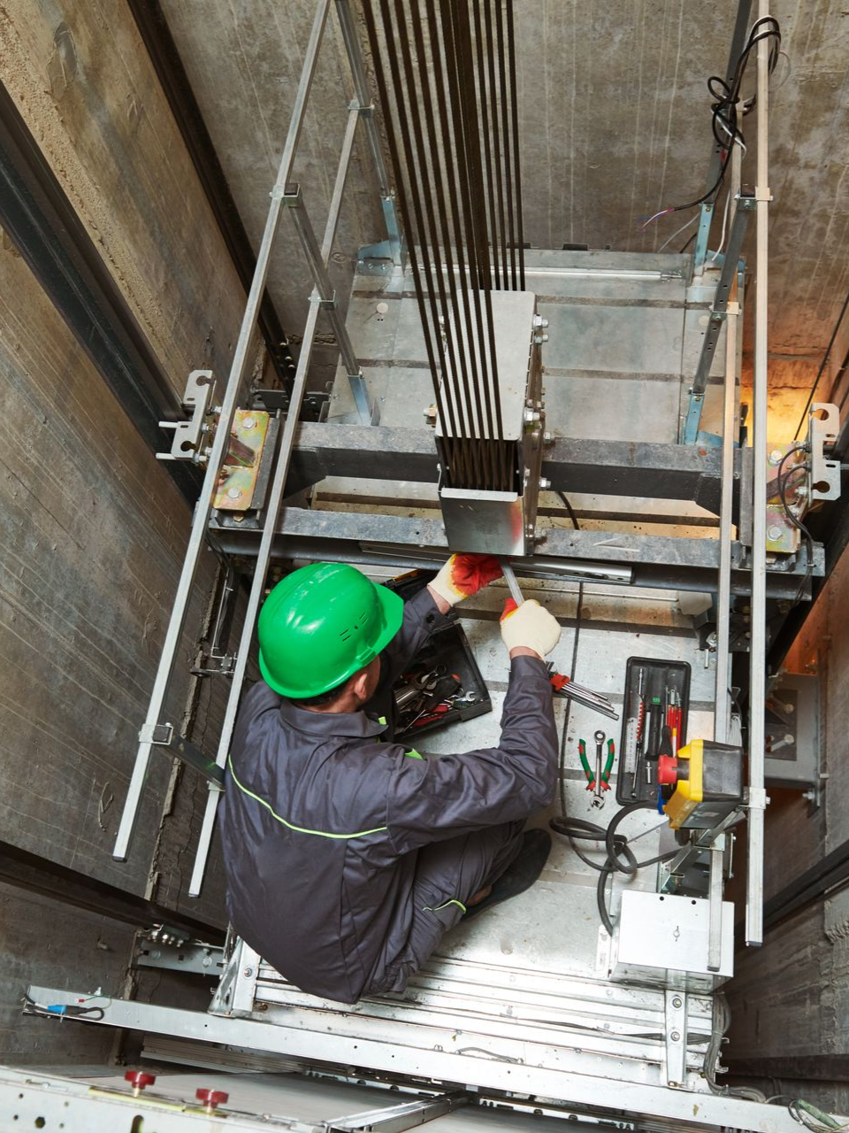 Une personne portant un casque vert travaille à l’intérieur d’une cage d’ascenseur, entourée de câbles et d’outils.